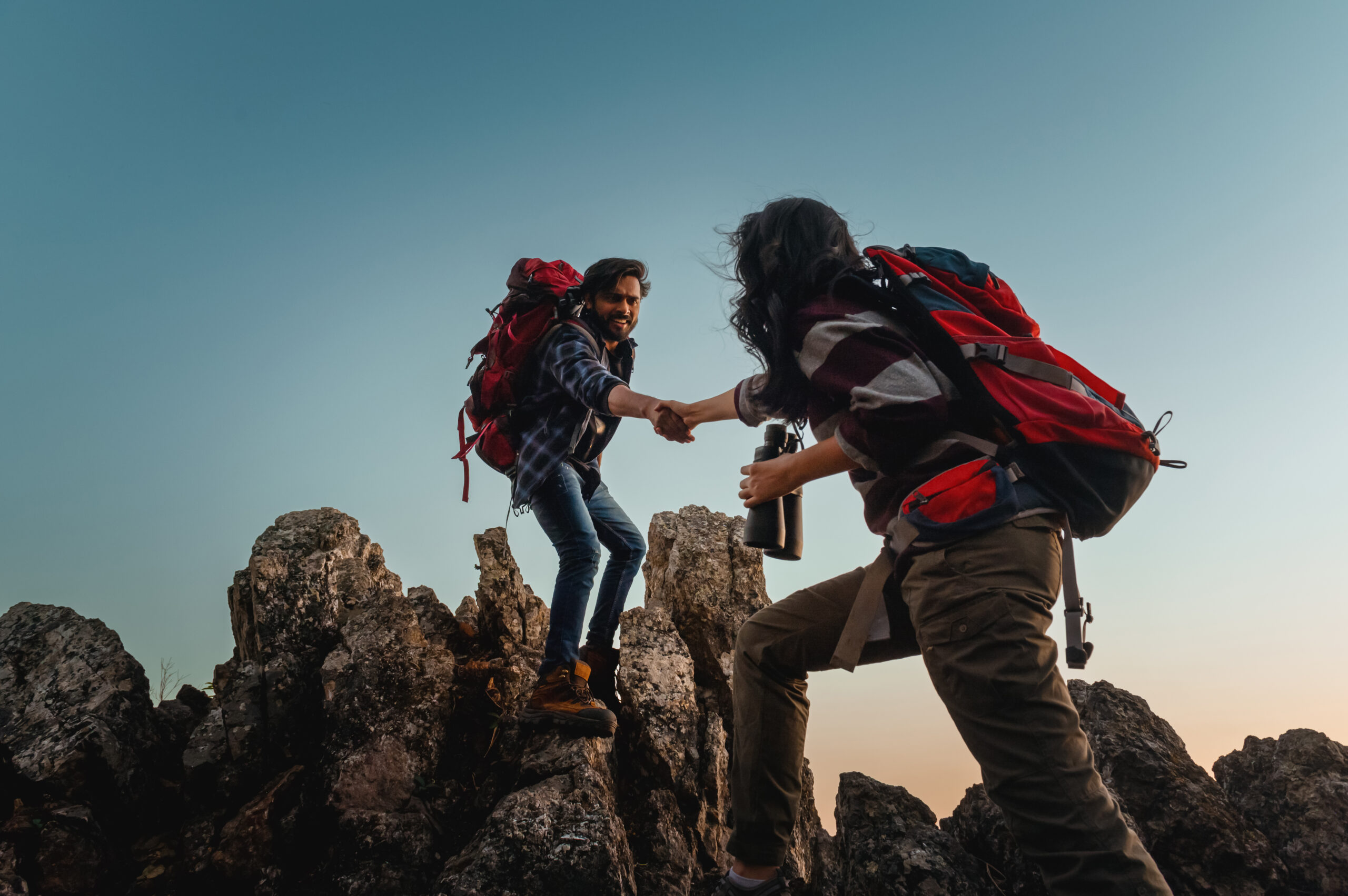 
Dos personas se ayudan mutuamente a escalar una montaña. Una de ellas lleva una mochila. La escena es tranquila y serena, con el sol brillando intensamente al fondo.
